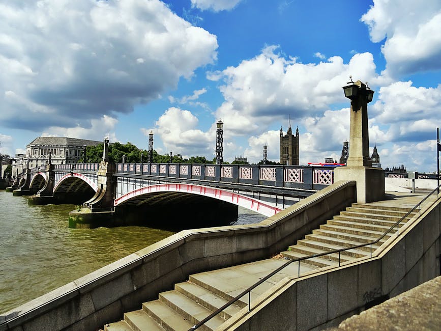 A view of Putney Bridge spanning the River Thames in London, with its distinctive white and red-painted arches supported by stone piers emerging from the water. Behind the bridge, there is a large, cl
