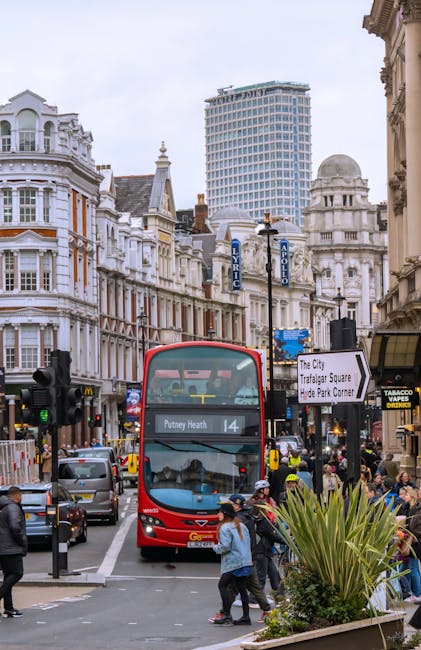 A busy urban street scene in Putney with a red double-decker bus approaching the intersection, displaying the route number 14 and destination Putney Heath on the front. Surrounding the bus are multiple vehicles, including cars and taxis, as well as pedestrians crossing the road. On the right side, people stand on the sidewalk near a small shop with a yellow sign and a tobacco products display. In the foreground, several individuals are walking, some carrying bags or backpacks, and others waiting to cross. The street is lined with classic Victorian-style terraced buildings featuring white facades, decorative architectural elements, and large windows. A tall modern building is visible in the background, contrasting with the older structures. Street signs indicate the location as Putney, with one pointing to Trafalgar Square and Hyde Park Corner. Bright daylight and overcast sky contribute to the lively atmosphere, complemented by streetlamps and greenery in planters, demonstrating a typical busy day in this part of London, with house removals and moving logistics activity often centered around such prominent locations.