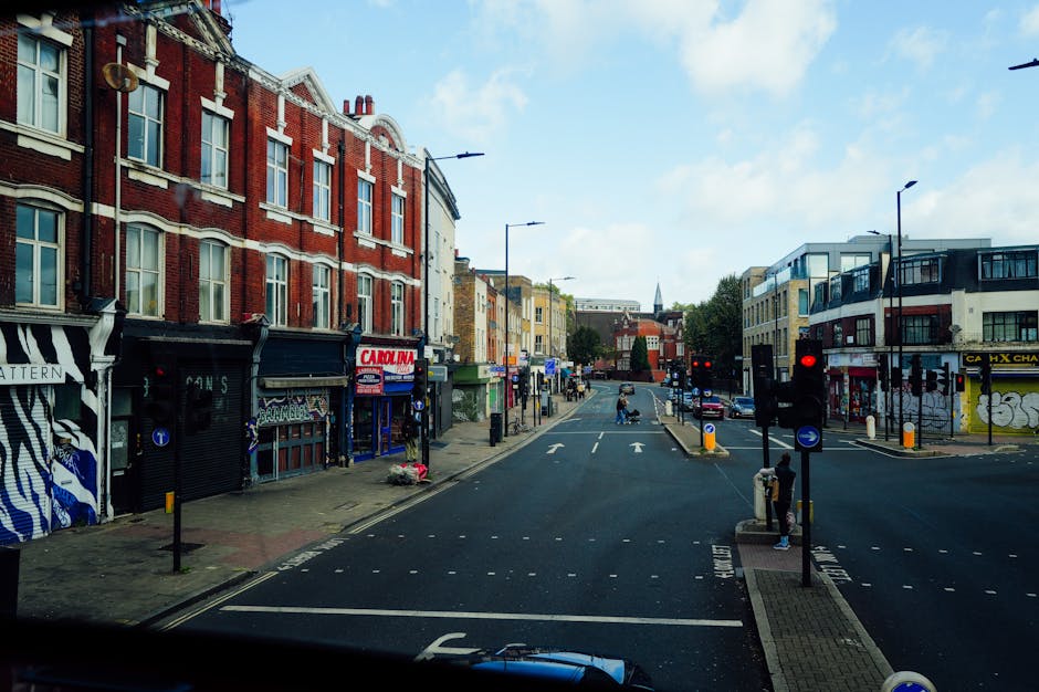 A view of a busy street scene in Putney, showing a row of mixed architectural style shops and residential buildings along the left side, some with canopy coverings and signage for local businesses. The street has several parked cars on both sides and is marked with white arrows indicating two-way traffic. Traffic lights are visible at the intersection, with one pedestrian waiting at the curb on the right. In the foreground, a person is seen standing near a small loading area with a few cardboard boxes and a hand trolley, suggesting preparations for a house or furniture removal. The scene is illuminated by natural daylight with a partly cloudy sky, and a van with the logo of Removal Companies Putney is positioned at the bottom edge, implying an ongoing home relocation or furniture transport process as part of professional removals services in the area.