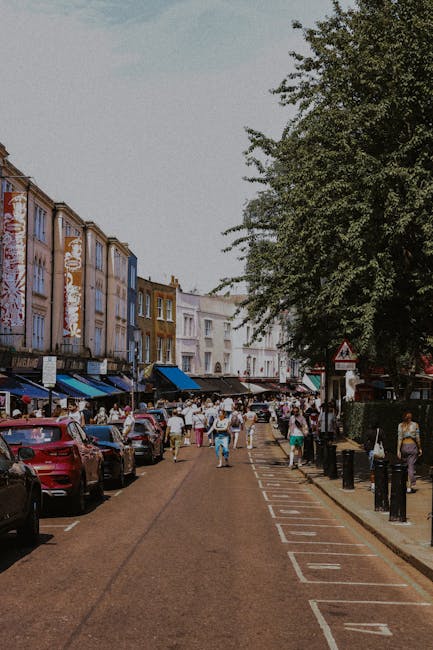 A busy urban street scene in Putney with a red double-decker bus approaching the intersection, displaying the route number 14 and destination Putney Heath on the front. Surrounding the bus are multiple vehicles, including cars and taxis, as well as pedestrians crossing the road. On the right side, people stand on the sidewalk near a small shop with a yellow sign and a tobacco products display. In the foreground, several individuals are walking, some carrying bags or backpacks, and others waiting to cross. The street is lined with classic Victorian-style terraced buildings featuring white facades, decorative architectural elements, and large windows. A tall modern building is visible in the background, contrasting with the older structures. Street signs indicate the location as Putney, with one pointing to Trafalgar Square and Hyde Park Corner. Bright daylight and overcast sky contribute to the lively atmosphere, complemented by streetlamps and greenery in planters, demonstrating a typical busy day in this part of London, with house removals and moving logistics activity often centered around such prominent locations.